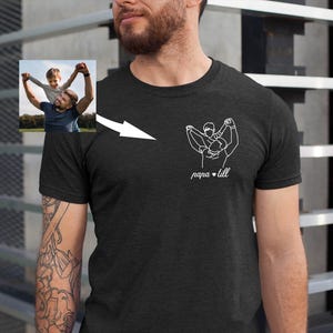 May include: Dark gray t-shirt featuring a white line drawing of a person holding a child, with the text "papa & till" below. A small photo of a man and child is in the upper left corner. The shirt is made of a soft material.