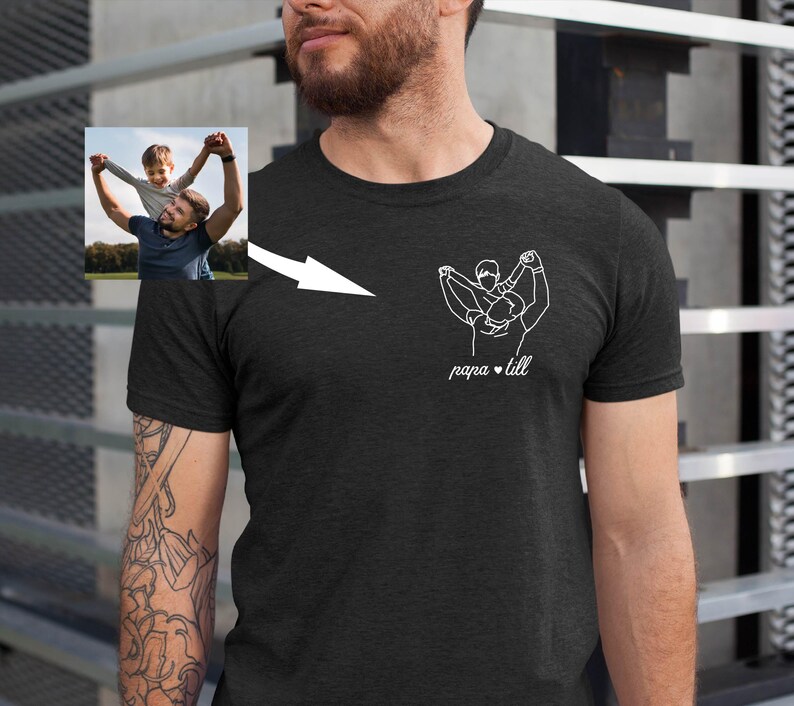 May include: Dark gray t-shirt featuring a white line drawing of a person holding a child, with the text "papa & till" below. A small photo of a man and child is in the upper left corner. The shirt is made of a soft material.