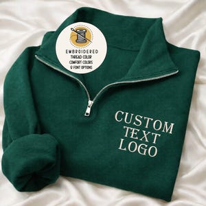 May include: A dark green quarter-zip sweatshirt with a silver zipper and white embroidered text that reads "CUSTOM TEXT LOGO." The sweatshirt has a round graphic with text that says "EMBROIDERED THREAD COLOR COMFORT COLORS & FONT OPTIONS."