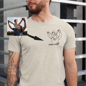 May include: A light gray t-shirt featuring a line drawing of a person holding a child, with the text "papa & lill" below. A small photo of a person holding a child is in the upper left corner, with a black arrow pointing to the line drawing.