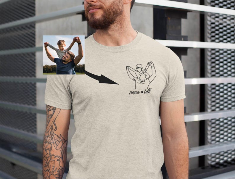 May include: A light gray t-shirt featuring a line drawing of a person holding a child, with the text "papa & lill" below. A small photo of a person holding a child is in the upper left corner, with a black arrow pointing to the line drawing.