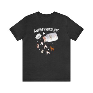 May include: Dark gray t-shirt featuring a graphic of a pill bottle labeled "ANTIDEPRESSANTS" with cartoon dogs spilling out. The design includes various dog breeds and small heart shapes, creating a playful and whimsical aesthetic.
