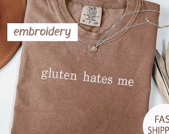 May include: Espresso-colored t-shirt with the embroidered text "gluten hates me." The shirt has a crew neck and short sleeves. A silver necklace with a small pendant rests on the shirt. The word "embroidery" is in a white box.