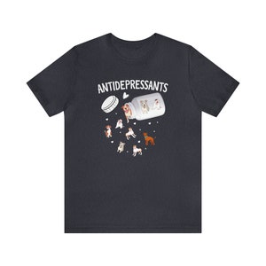 May include: Dark gray t-shirt with the word "ANTIDEPRESSANTS" above a graphic of a pill bottle spilling out cartoon dogs. The dogs are various breeds and colors, with hearts scattered around them.