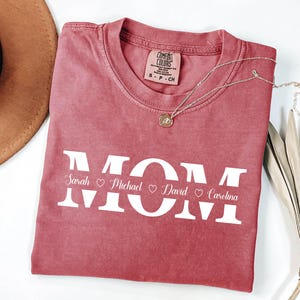 May include: A red t-shirt with the word "MOM" in white letters. The names "Sarah", "Michael", "David", and "Carolina" are written below the letters with a heart symbol between each name.