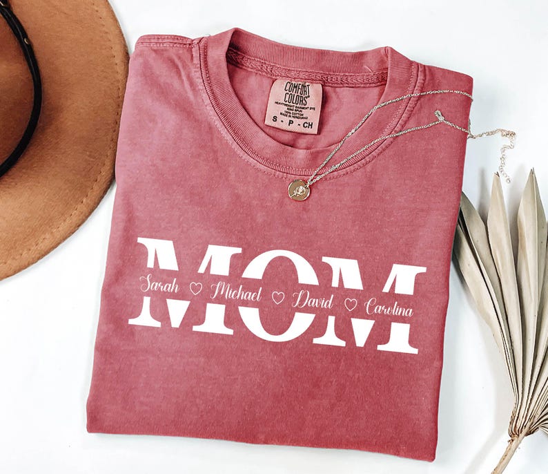 May include: A red t-shirt with the word "MOM" in white letters. The names "Sarah", "Michael", "David", and "Carolina" are written below the letters with a heart symbol between each name.