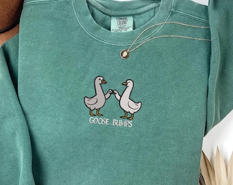 May include: A teal Comfort Colors sweatshirt with an embroidered design of two geese holding hands. The text "GOOSE BUMPS" is embroidered below the geese. A gold necklace with a pendant is draped on the sweatshirt.