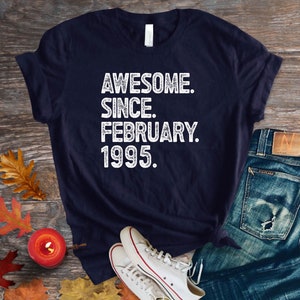 May include: Navy blue t-shirt with the text "AWESOME. SINCE. FEBRUARY. 1995." in white. The shirt is paired with blue jeans and white sneakers on a wooden surface. Autumn leaves and a candle are also present.