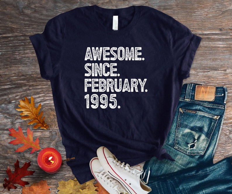 May include: Navy blue t-shirt with the text "AWESOME. SINCE. FEBRUARY. 1995." in white. The shirt is paired with blue jeans and white sneakers on a wooden surface. Autumn leaves and a candle are also present.