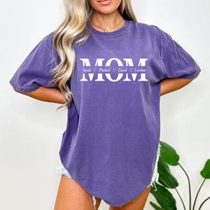 May include: A purple t-shirt with the word "MOM" printed in white. Below the word "MOM" are the names "Sarah", "Michael", "David", and "Carolina" printed in a smaller font.