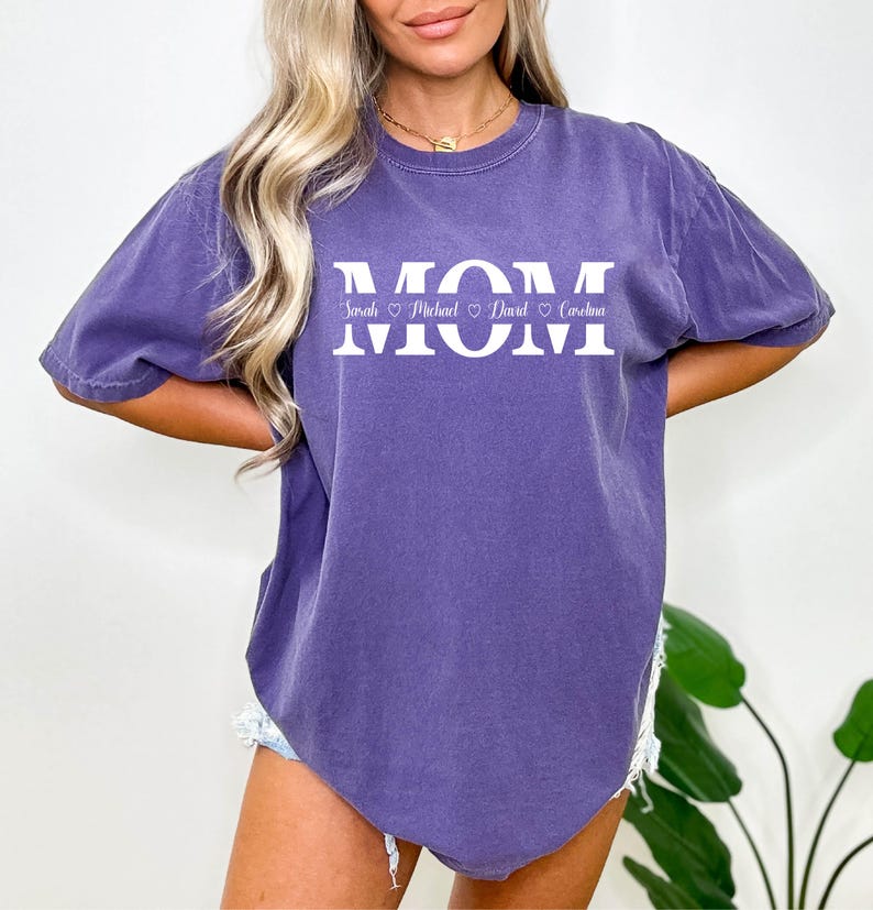 May include: A purple t-shirt with the word "MOM" printed in white. Below the word "MOM" are the names "Sarah", "Michael", "David", and "Carolina" printed in a smaller font.