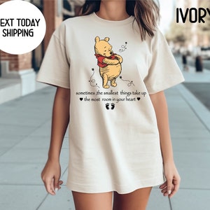 Peut inclure: Un t-shirt beige avec une image de dessin anim&eacute; de Winnie l'ourson en train d'embrasser un pot de miel. Le texte "sometimes, the smallest things take up ❤ the most room in your heart ❤" est imprim&eacute; sous l'image.