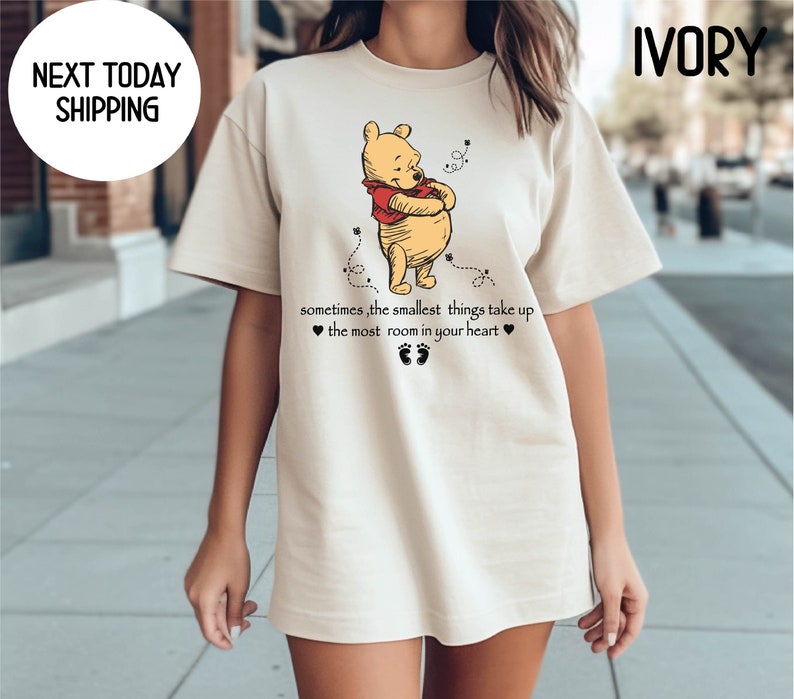Peut inclure: Un t-shirt beige avec une image de dessin anim&eacute; de Winnie l'ourson en train d'embrasser un pot de miel. Le texte "sometimes, the smallest things take up ❤ the most room in your heart ❤" est imprim&eacute; sous l'image.