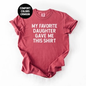 May include: Crimson red t-shirt with the text "MY FAVORITE DAUGHTER GAVE ME THIS SHIRT" in white block letters. The shirt has rolled-up sleeves and a crew neck. A black circle in the upper left corner reads "COMFORT COLORS CRIMSON".