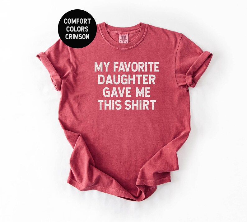 May include: Crimson red t-shirt with the text "MY FAVORITE DAUGHTER GAVE ME THIS SHIRT" in white block letters. The shirt has rolled-up sleeves and a crew neck. A black circle in the upper left corner reads "COMFORT COLORS CRIMSON".