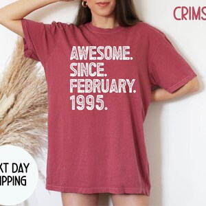 May include: Crimson-colored t-shirt with the text "AWESOME. SINCE. FEBRUARY. 1995." in white. The shirt has short sleeves and a crew neck. The words "NEXT DAY SHIPPING" are also visible.