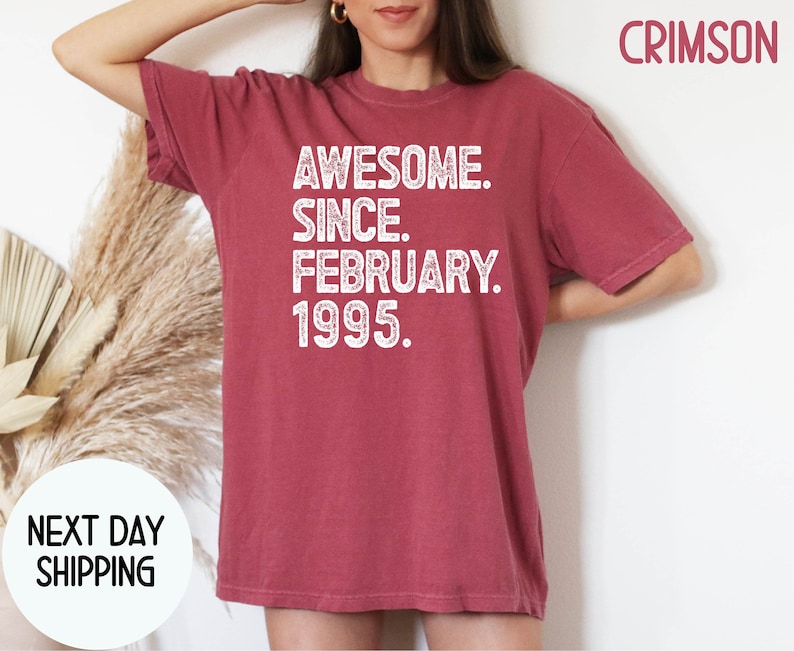May include: Crimson-colored t-shirt with the text "AWESOME. SINCE. FEBRUARY. 1995." in white. The shirt has short sleeves and a crew neck. The words "NEXT DAY SHIPPING" are also visible.