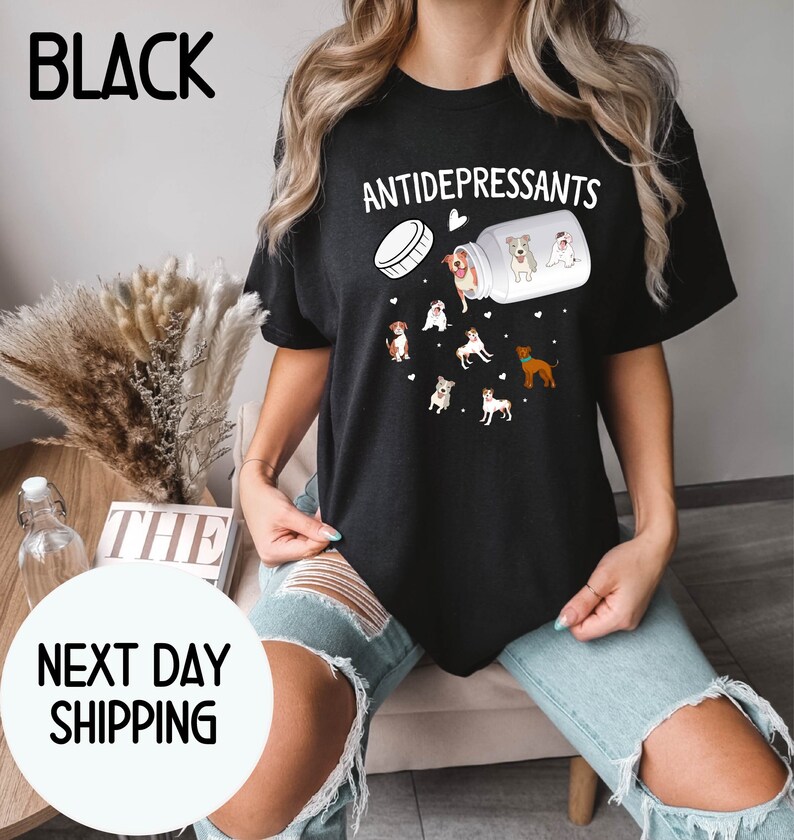 May include: Black t-shirt with the word "ANTIDEPRESSANTS" above a graphic of a pill bottle spilling out cartoon dogs. The shirt also has the word "BLACK" printed at the top left.