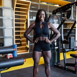 Könnte beinhalten: Eine Frau mit langen schwarzen Zöpfen lächelt, während sie in einem Fitnessstudio steht. Sie trägt einen schwarzen Sport-BH, schwarze Shorts und weiße Turnschuhe. Sie steht vor einem Gewichtsgerät.