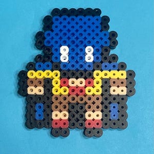 May include: Pixel art figure made from colorful plastic beads. The figure has a blue head, white eyes, and a body with yellow, brown, red, and black details. The background is a solid light blue.