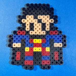 May include: A pixel art rendition of Superman, made with colorful beads. The figure is wearing a red and blue suit with a yellow 'S' symbol on the chest. The figure has black hair and a black cape.