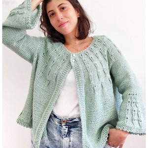 Könnte beinhalten: Hellgrüne Strickjacke mit weiten Ärmeln und strukturiertem Muster. Der offene Cardigan wird mit zwei Knöpfen geschlossen und hat eine gewellte Bordüre an den Ärmeln und Kanten. Getragen über einem weißen Oberteil und blauen Jeans.