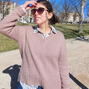 May include: A woman wearing a pink knitted sweater with a v-neck and a blue and white plaid shirt underneath. She is wearing sunglasses with a beaded chain.