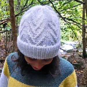 May include: A white knitted beanie with a cable knit pattern. The beanie is being worn by a person with dark hair and a blue and yellow striped jumper.