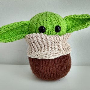 Puede incluir: Un juguete de peluche verde tejido con orejas grandes y ojos negros, que lleva una bufanda de punto de color crema. El juguete es marrón en la parte inferior.