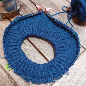 May include: A dark blue knitted circle with multiple stitch markers attached to the edge. The circle is being worked on with knitting needles and yarn.
