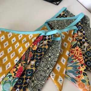 May include: A selection of fabric bunting flags in various patterns. The designs include yellow with white diamonds, floral patterns, and a grey floral print. Each flag is trimmed with a light blue border.