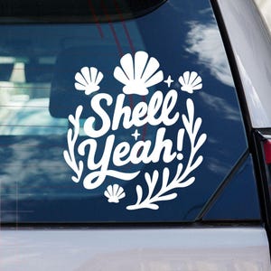 May include: A white vinyl decal applied to a car window featuring the text Shell Yeah! in a cursive script font, surrounded by illustrations of four seashells and two leafy branches, with small four-pointed star accents scattered throughout.