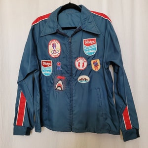 May include: A blue vintage jacket with red and white trim. The jacket has several patches, including "Montreal Olympics 76", "Dlistex", "Jaws", and "Master 76".