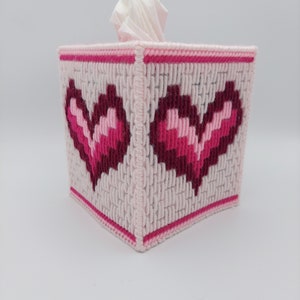 May include: Pink and red heart-shaped tissue box cover made with yarn. The cover is square and has a white tissue box inside.