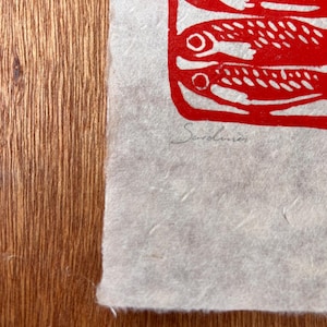 May include: A textured off-white paper print featuring a bold red ink illustration of stacked fish. The word Sardines is handwritten in pencil below the red graphic. The paper has a deckled edge and a visible fibrous, handmade quality throughout.