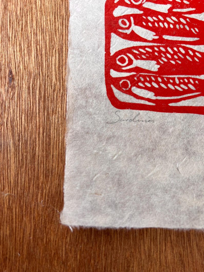 May include: A textured off-white paper print featuring a bold red ink illustration of stacked fish. The word Sardines is handwritten in pencil below the red graphic. The paper has a deckled edge and a visible fibrous, handmade quality throughout.
