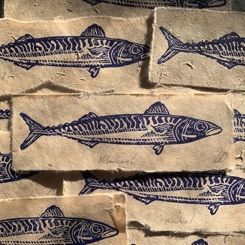 May include: A collection of rectangular handmade paper sheets featuring a dark blue ink block print of a mackerel fish. Each print displays the fish in profile with detailed line work and the handwritten word Mackerel in cursive script.