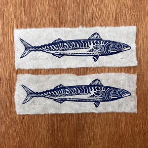 May include: Two rectangular pieces of textured off-white paper, each featuring a dark blue ink block print of a mackerel fish. The fish illustration displays detailed scales and fins, with the word Mackerel written in pencil below the fish.