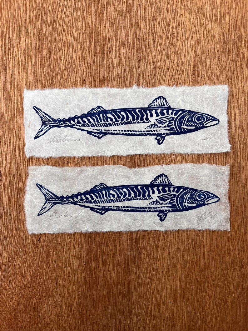 May include: Two rectangular pieces of textured off-white paper, each featuring a dark blue ink block print of a mackerel fish. The fish illustration displays detailed scales and fins, with the word Mackerel written in pencil below the fish.