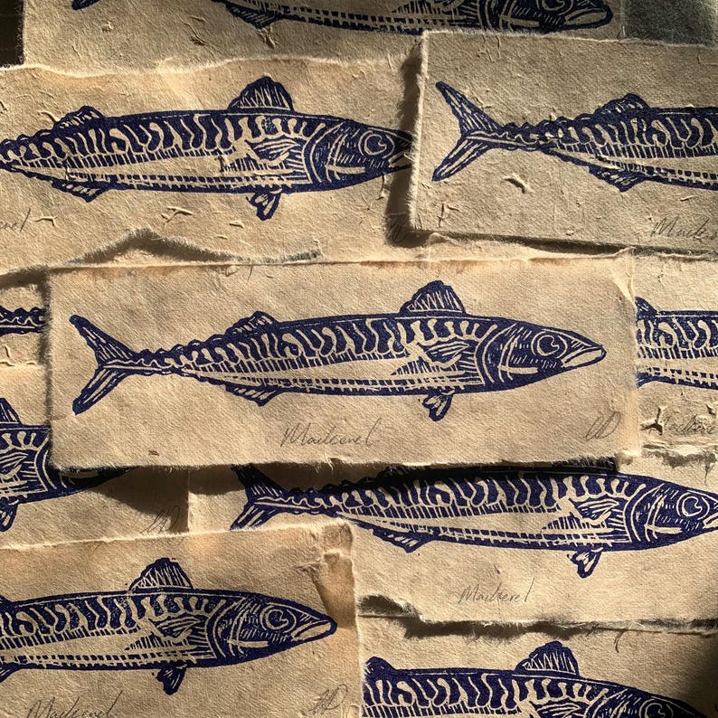 May include: A collection of rectangular paper scraps featuring dark blue ink block prints of mackerel fish. Each piece of textured, off-white paper displays a single fish illustration with the handwritten word Mackerel below the image.