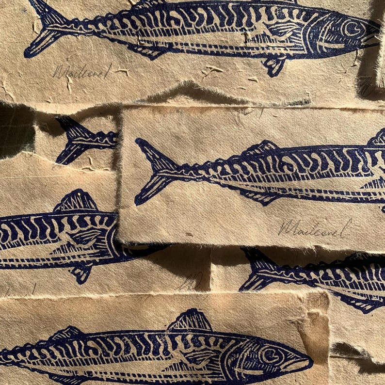 May include: Multiple overlapping rectangular pieces of light tan textured paper, each featuring a dark blue ink block print of a mackerel fish. The word Mackerel is handwritten in pencil below each fish illustration on the paper strips.