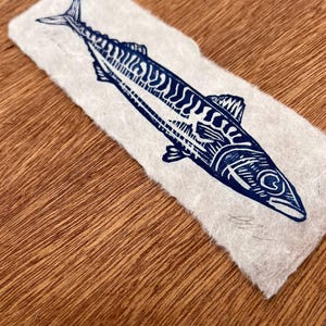 May include: A rectangular piece of off-white, textured paper featuring a dark blue ink block print of a mackerel fish. The fish is depicted in profile with detailed line work showing its scales and fins. A handwritten signature is visible.