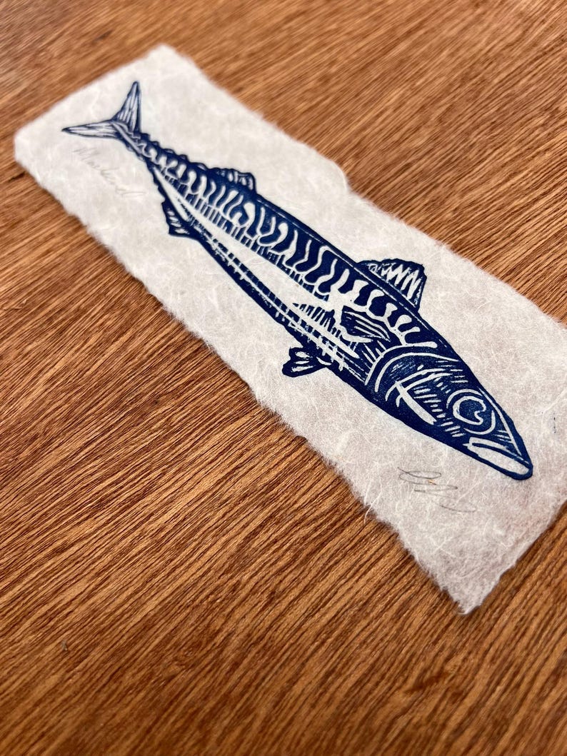 May include: A rectangular piece of off-white, textured paper featuring a dark blue ink block print of a mackerel fish. The fish is depicted in profile with detailed line work showing its scales and fins. A handwritten signature is visible.