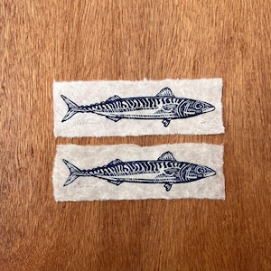May include: Two rectangular pieces of off-white textured paper, each featuring a dark blue ink block print of a mackerel fish. The fish illustrations show detailed scales and fins, with the word Mackerel written in pencil below each fish.