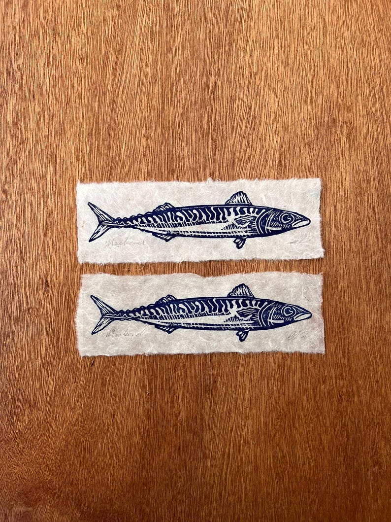 May include: Two rectangular pieces of off-white textured paper, each featuring a dark blue ink block print of a mackerel fish. The fish illustrations show detailed scales and fins, with the word Mackerel written in pencil below each fish.