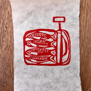 May include: A rectangular piece of textured, off-white paper featuring a red ink block print of a tin of sardines. The illustration depicts four fish stacked horizontally inside a container with a key opener. The word Sardines is written in pencil.