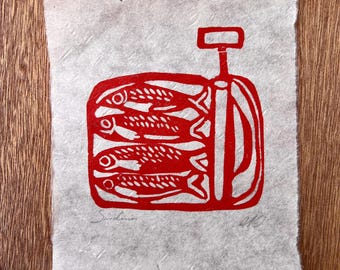 May include: A rectangular piece of textured, off-white paper featuring a red ink block print of a tin of sardines. The illustration depicts four fish stacked horizontally inside a container with a key opener. The word Sardines is written in pencil.