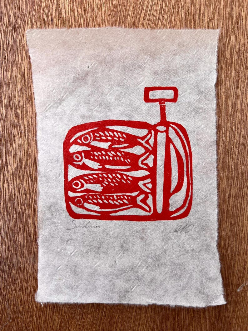 May include: A rectangular piece of textured, off-white paper featuring a red ink block print of a tin of sardines. The illustration depicts four fish stacked horizontally inside a container with a key opener. The word Sardines is written in pencil.