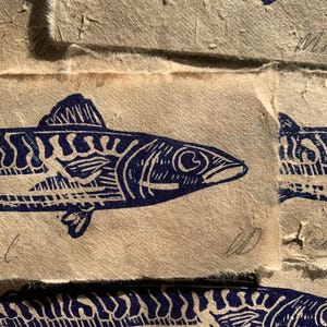 May include: A dark blue ink block print of a fish on textured, light tan handmade paper. The fish features detailed line work representing scales and fins. A handwritten signature is visible in the bottom right corner of the paper print.