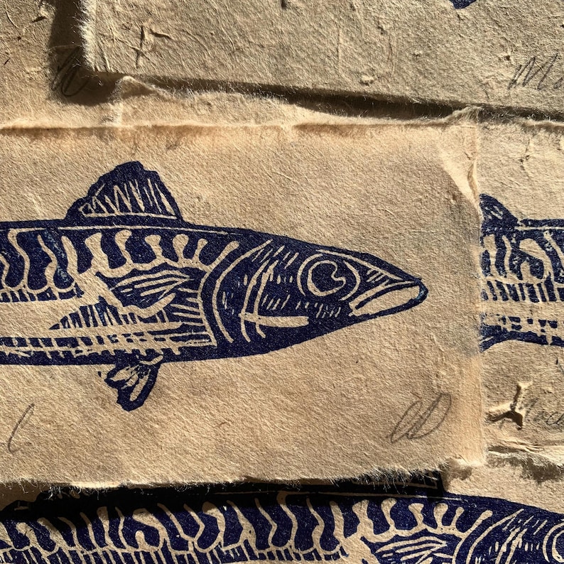 May include: A dark blue ink block print of a fish on textured, light tan handmade paper. The fish features detailed line work representing scales and fins. A handwritten signature is visible in the bottom right corner of the paper print.